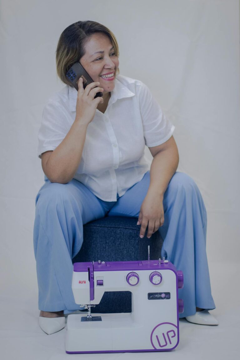 Cheerful woman in casual attire talking on phone, sitting by sewing machine.